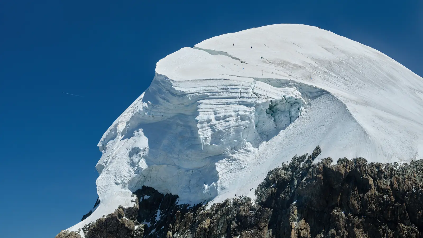 The Breithorn’s summit ice cap from the air: a dome of snow ending in broken ice cliffs above a band of dark rock.