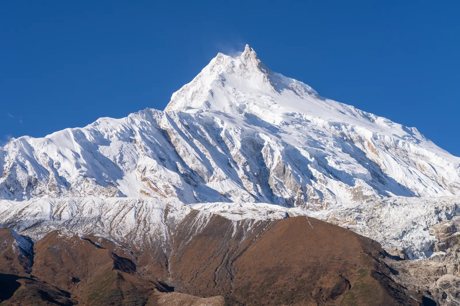 Manaslu’s summit ridge and fluted snow faces above brown foothills, under a clear sky.