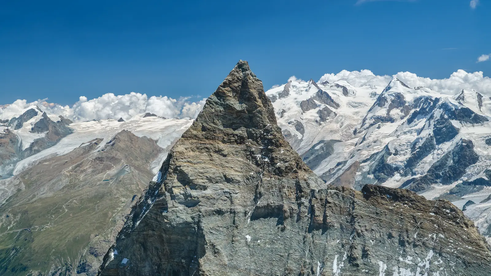 The Matterhorn from the air: a steep pyramid of rock and snow above glaciers, with a line of snow peaks and cloud behind.